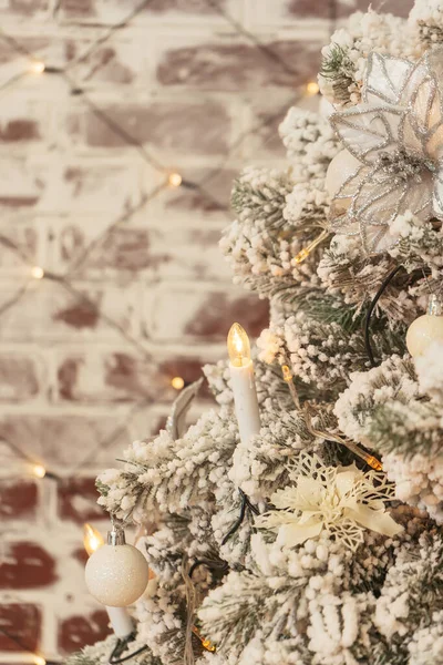 Garlands in the shape of a candle on a snow-covered Christmas tree. White silver ornaments poinsettia balls snowflakes on the Christmas tree. Christmas tree with snow on the background of a brick wall close up