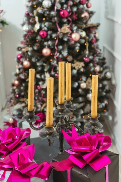 Golden candles in a candelabrum on a table with gifts boxes on a background of the Christmas tree. Gift boxes in black and silver wrapping paper with pink bows with ribbons on a white wall with stucco molding. Stylish Christmas home decor
