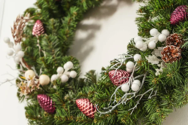 白い果実とピンクのバーガンディプラスチック光沢のあるコーンクリスマスの装飾が壁に偽のトウヒの花輪 詳細ホームスタジオのクリスマスインテリア 白い背景のクリスマスの花輪クローズアップ — ストック写真