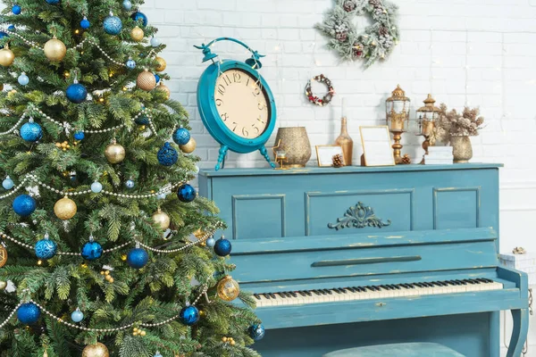 Blue piano with Christmas decor wreaths on the wall. Large clock alarm clock on piano new year decor. The room is decorated in a classic style with a piano and a Christmas tree in blue and gold style