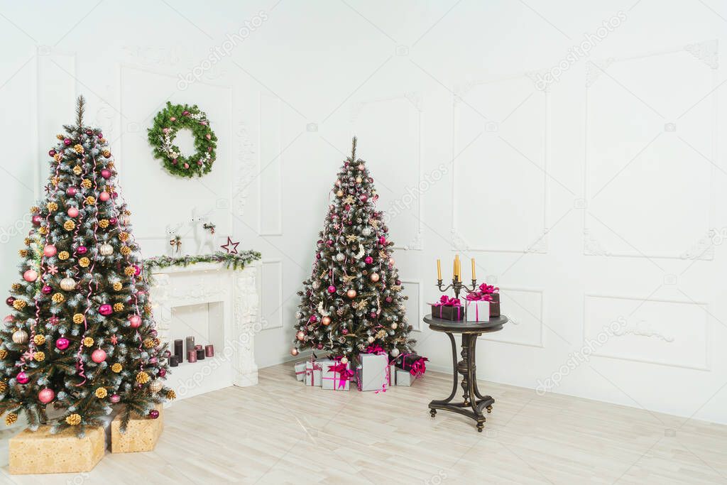 White fireplace with stucco in the classic Christmas decoration of the living room. Balloons ribbon garlands in pink gold maroon style on fake Christmas tree decorations. Christmas trees with cones in gold silver glitter sequins.
