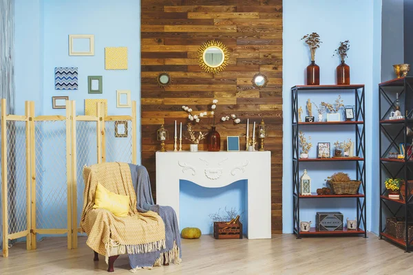 A beige armchair with a yellow blanket and a fake fireplace in a room with blue walls. Macrame decoration and cotton twigs in a bottle in a cozy living room. Wooden wall above fireplace in interior