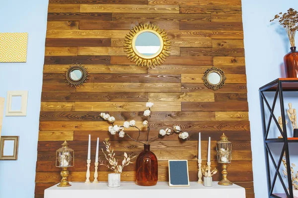 A wooden wall over a white fireplace. White classic false fireplace with autumn decorations. Cotton branches in a bottle in the interior. Sun-shaped mirrors on the wall