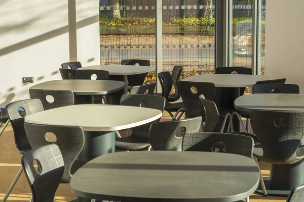 Modern masa ve sandalyelerle dolu boş bir kantinin içi gün ışığında geniş pencerelerden aydınlatılıyor.