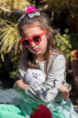 Üzgün yüzlü, elinde oyuncakla bahçede bankta oturan beyaz bir kız. Açık havada bir oyuncakla çocuk portresi..