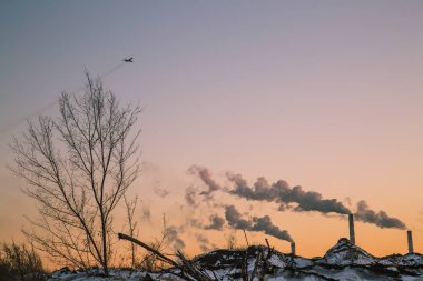 Uçak iniyor, fabrikanın tüten borularının üzerinden uçuyor güzel bir kış günbatımının arka planında.