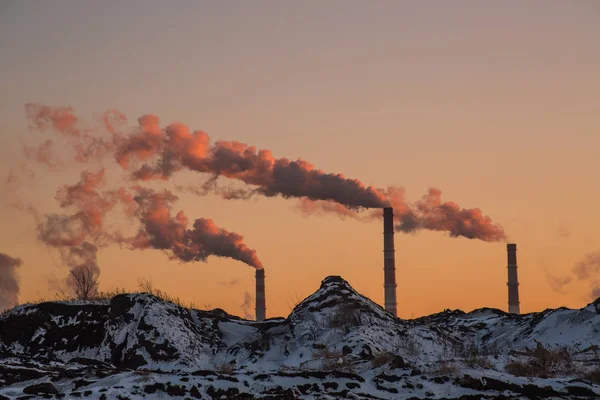 the smoky chimneys of the factory rise above the mountains of earth at sunset