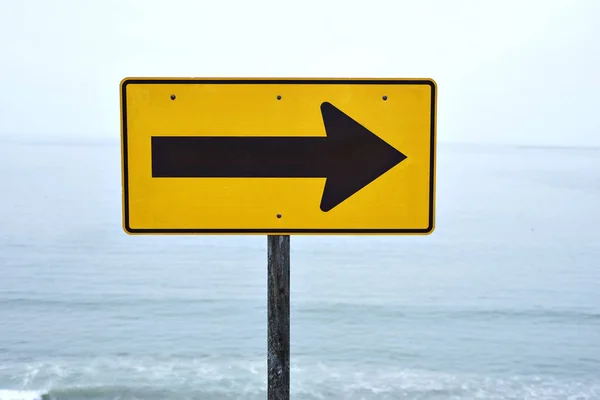 A yellow street sign with arrow pointing right at the beach with a view of the ocean