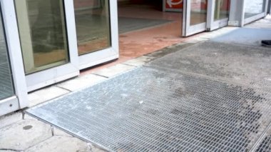 Otomatik cam kapılar açılıp kapanıyor. Alışveriş merkezine giriş. Kış. Yerde ıslak ayak izleri var. Yoldan geçenlerin bacakları.