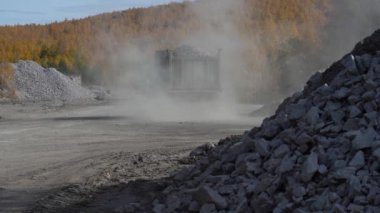 Dolu bir damperli kamyon, açık güneşli bir günde, sarı ağaçların arasında tozlu bir yol boyunca ilerler. Sonbaharda bir orman yolunda taşlarla dolu bir çöp kamyonu geziyor..