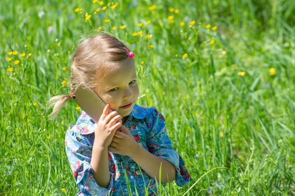 Cute little girl is talking on the phone. A little girl sits with a mobile phone in a green meadow on a sunny summer day. The serious face of a child talking on the phone — Stockfoto