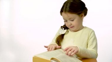 A child and a book in the studio on a white background. Little girl smiling and talking, leafing through a thick book. A child of 5-6 years old is looking for pictures in a book