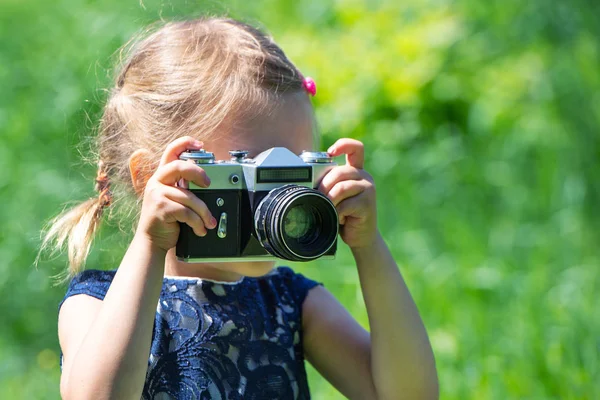 Küçük bir kız, yazın parkta retro film kamerasıyla fotoğraf çekiyor. Açık renk elbiseli, elinde yeşil çimlerin üzerinde antika fotoğraf makinesi olan küçük tatlı bir kız bebek..