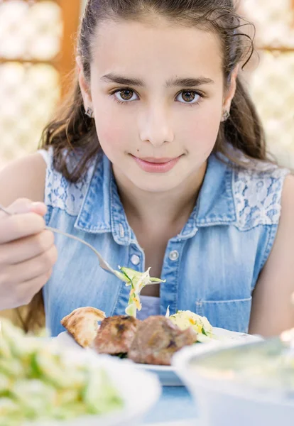 Маленькая красивая улыбающаяся девочка с удовольствием ест шашлык на открытом воздухе — стоковое фото