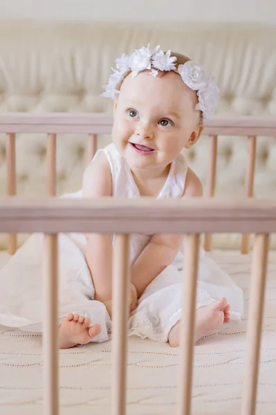 A happy baby in a dress sits in a crib — Stock Photo, Image