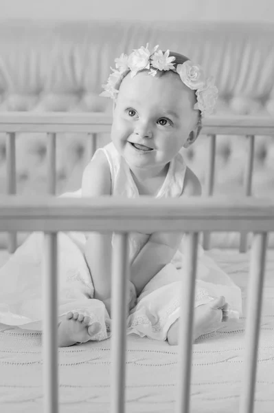 A happy baby in a dress sits in a crib — Stock Photo, Image