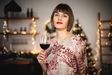 Uzun elbiseli, kendine güvenen bir kızın, bir bardak şarapla Noel ağacının yanında poz verdiği bir resim..
