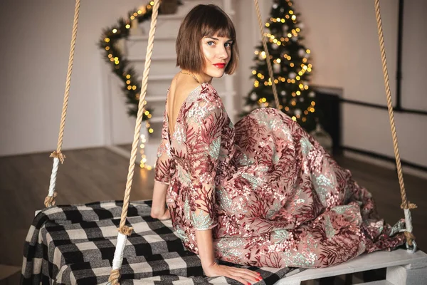 Uzun elbiseli, kendine güvenen bir kızın salıncaktaki Noel ağacının yanında poz verdiği bir resim..