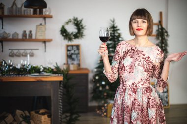 Uzun elbiseli, kendine güvenen bir kızın, bir bardak şarapla Noel ağacının yanında poz verdiği bir resim..