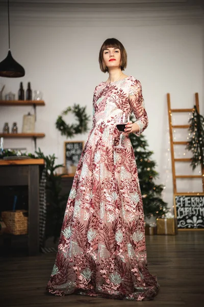 Indoor portrait of confident girl in a long dress posing near christmas tree with glass of wine. — Stock Photo, Image