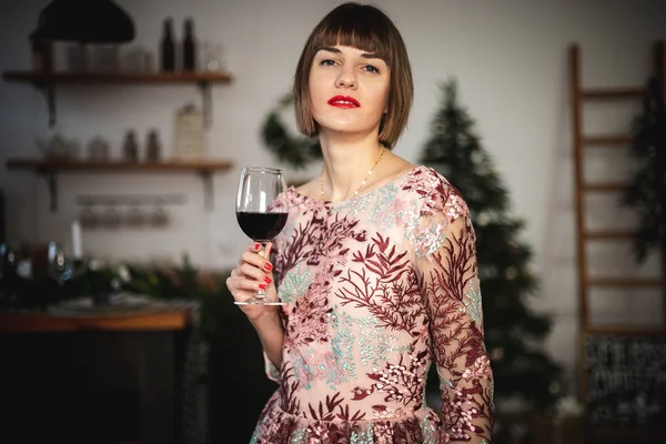 Uzun elbiseli, kendine güvenen bir kızın, bir bardak şarapla Noel ağacının yanında poz verdiği bir resim..