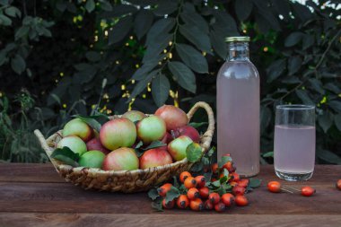 Bir cam şişenin içinde ve bir bardağın içinde olgun elma ve gül üzümleriyle dolu bir sepetin içinde yeşil yapraklardan oluşan bir arka planda duran ahşap bir masanın üzerinde duruyor.