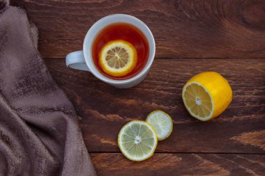 Dilimlenmiş beyaz bardak çay ve kahverengi ahşap bir masada yarım limon ve sol üst manzarasında kahverengi bir peçete.