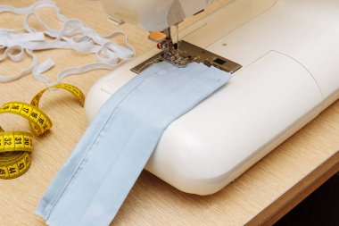 Mavi kumaştan yapılmış bitmemiş bir tıbbi maske beyaz bir dikiş makinesinin üzerinde ve yanında elastik bir bant ve bir santimetre uzunluğunda bir bant var.