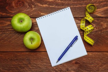 Bir yay boş beyaz çarşaflar ve yanında yeşil elmalar olan bir kilo kaybı planı için bir kalem ve kahverengi tahta masa görünümlü bir mezura ile dolu bir not defteri.