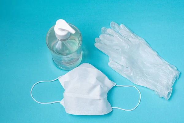 Açık mavi arka planda alkol jeli, eldiven ve cerrahi maske olan bir kap.