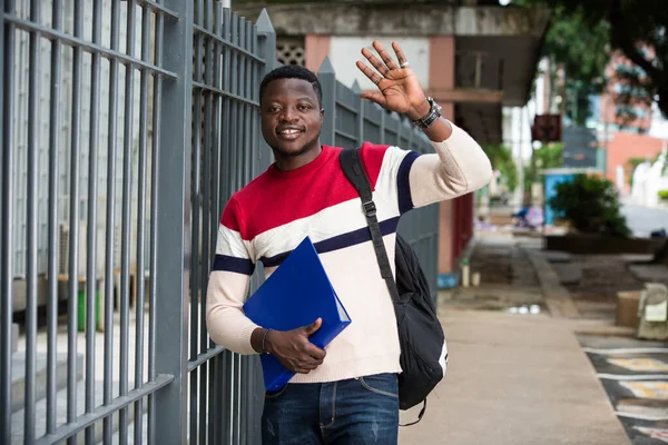 Young student with hand gesture in greeting or welcome background Man with beard and books in hand in the context of a university building.