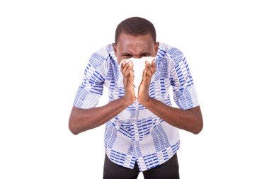 Gömlekli genç bir adam beyaz bir arka planda duruyor hapşırıyor ve ağzını mendille örtüyor..