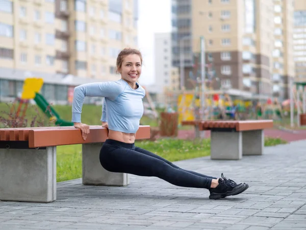Kaukázusi vörös hajú lány fordított fekvőtámaszt csinál egy padon a sportcsarnokban, szelektív fókusz. Kültéri napi edzés. — Stock Fotó