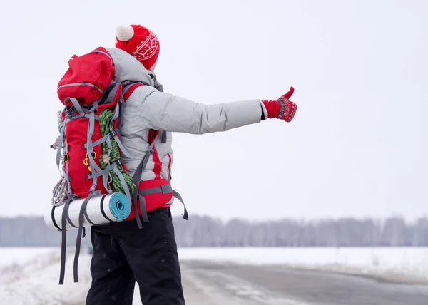 雪の田舎道に親指を持ち上げ大きな赤いバックパックで冬のスポーツウェアでヒッチハイカー、バックビュー、選択した焦点を共有する. — ストック写真