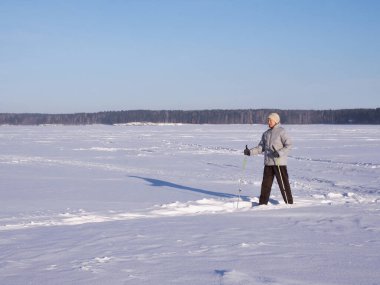 Yaşlı bir kadın kışın, güneşli bir günde, karla kaplı bir gölde, açık havada İskandinav yürüyüşü yapıyor..