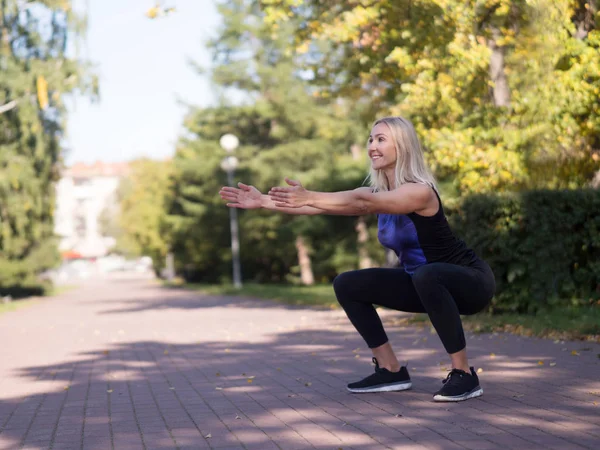 Biała uśmiechnięta sportowiec w średnim wieku kobieta robi głęboki squat na chodniku w miejskim parku, outdoor, selektywny ostrość — Zdjęcie stockowe