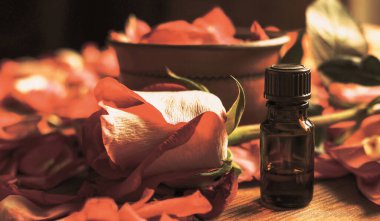 Ahşap masadaki gül yaprakları arasında koyu renk cam şişe ve gül goncası, tonlanmış sıcak, seçilmiş odak noktası.