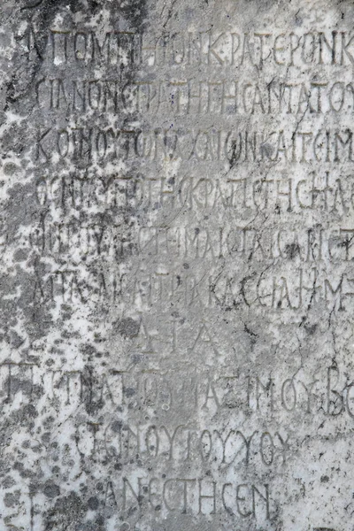 Yarısı silinmiş antik Yunan harfleri eski gri mermer bir levhaya işlenmiş çatlaklar ve kalıplarla kaplı..