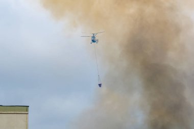 Moskova, Rusya - Ağustos 2019: İtfaiyeciler iş başında. Su dolu süspansiyon gondollu itfaiye helikopteri yangından çıkan siyah-turuncu dumandan çıkan büyük bir yangını söndürüyor..