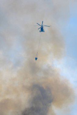 Moskova, Rusya - Ağustos 2019: İtfaiyeciler iş başında. Su dolu süspansiyon gondollu itfaiye helikopteri yangından çıkan siyah-turuncu dumandan çıkan büyük bir yangını söndürüyor..