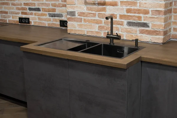 Modern kitchen with black sink, kitchen faucet for water and fronts against the background of a brick wal. Brown and black wooden countertops. Luxury dark themed background for interior.