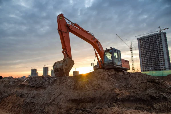 Excavator on earthworks at construction site on sunset background. Backhoe digs ground for the foundation and for paving out sewer line. Construction machinery for excavating