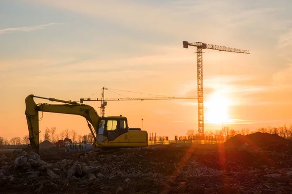 Excavator and tower crane at construction site on sunset background. Construction crane constructing a new building. Renovation program, development, concept of the buildings industry. Soft focus