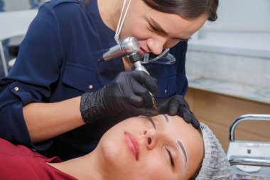 Usta, bir dövme makinesiyle müşteriye kaş makyajı yapar.