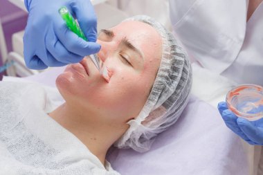 Masada yatan havlu genç kadının yüzünde ve boynunda fırça ile beyaz maske uygulayan kırpma ustası