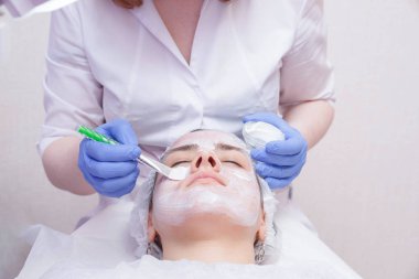 Masada yatan havlu genç kadının yüzünde ve boynunda fırça ile beyaz maske uygulayan kırpma ustası