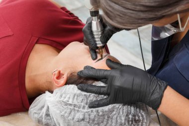 Usta, dövme makinesi kullanarak müşteriye kaş maskesi takıyor. Kalıcı Kaş Makyajı Prosedürü.