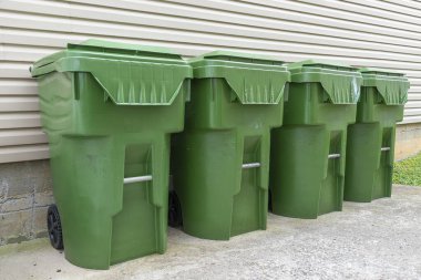Dört yeşil plastik çöp tenekesinin yatay görüntüsü bir apartmanın dış duvarına karşı dizilmiş..