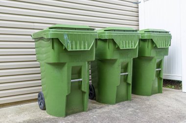 Üç yeşil plastik çöp tenekesinin yatay görüntüsü bir apartmanın dış duvarına karşı dizilmiş..