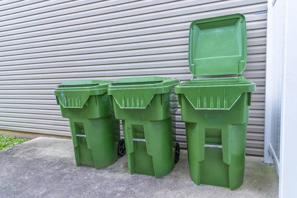 Üç yeşil plastik çöp tenekesinin yatay görüntüsü bir apartmanın dış duvarına karşı dizilmiş. Birinin kapağı açık..